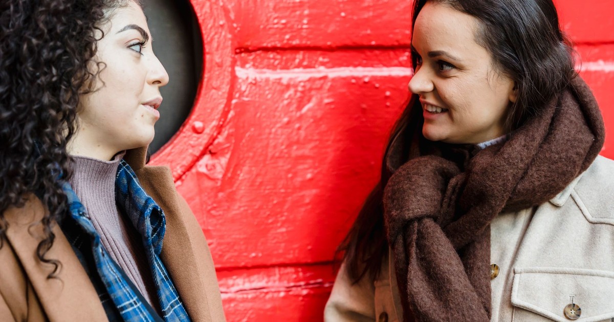 Positive young multiethnic female friends in casual outfits standing on street near red metal door and chatting
