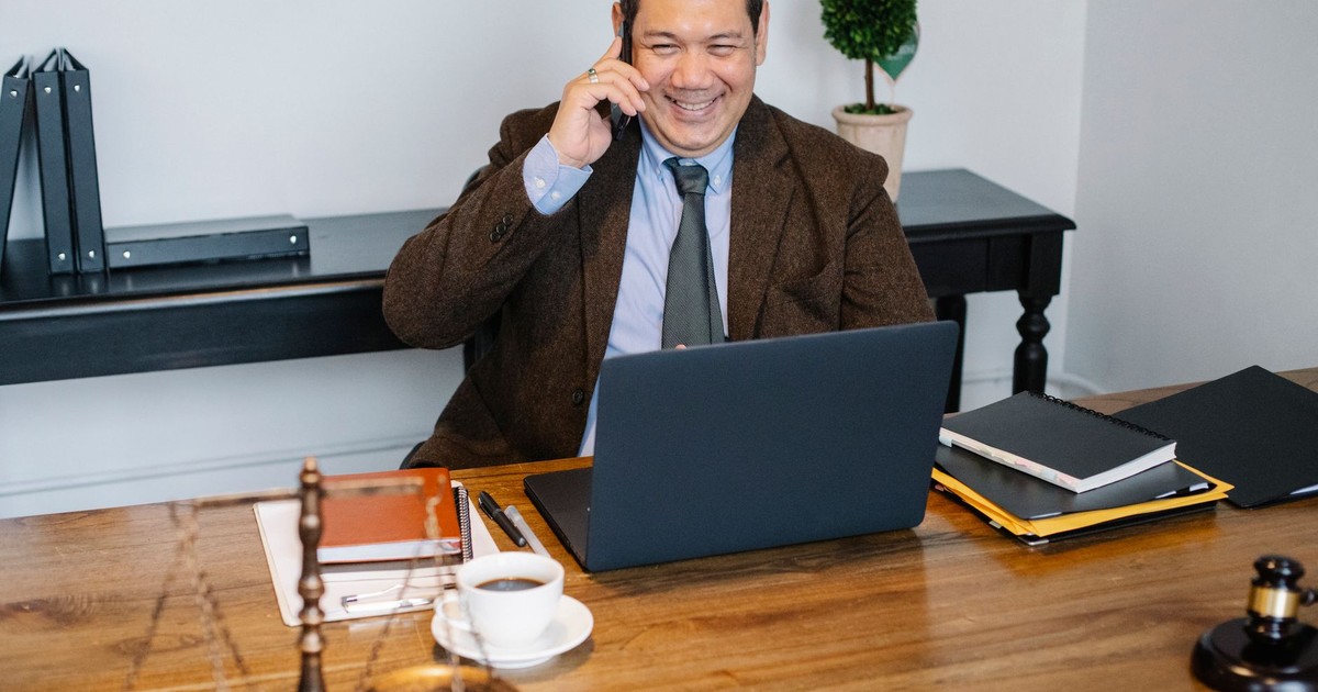 Joyful Asian male lawyer wearing formal suit having conversation on mobile phone and using laptop while working in modern law firm