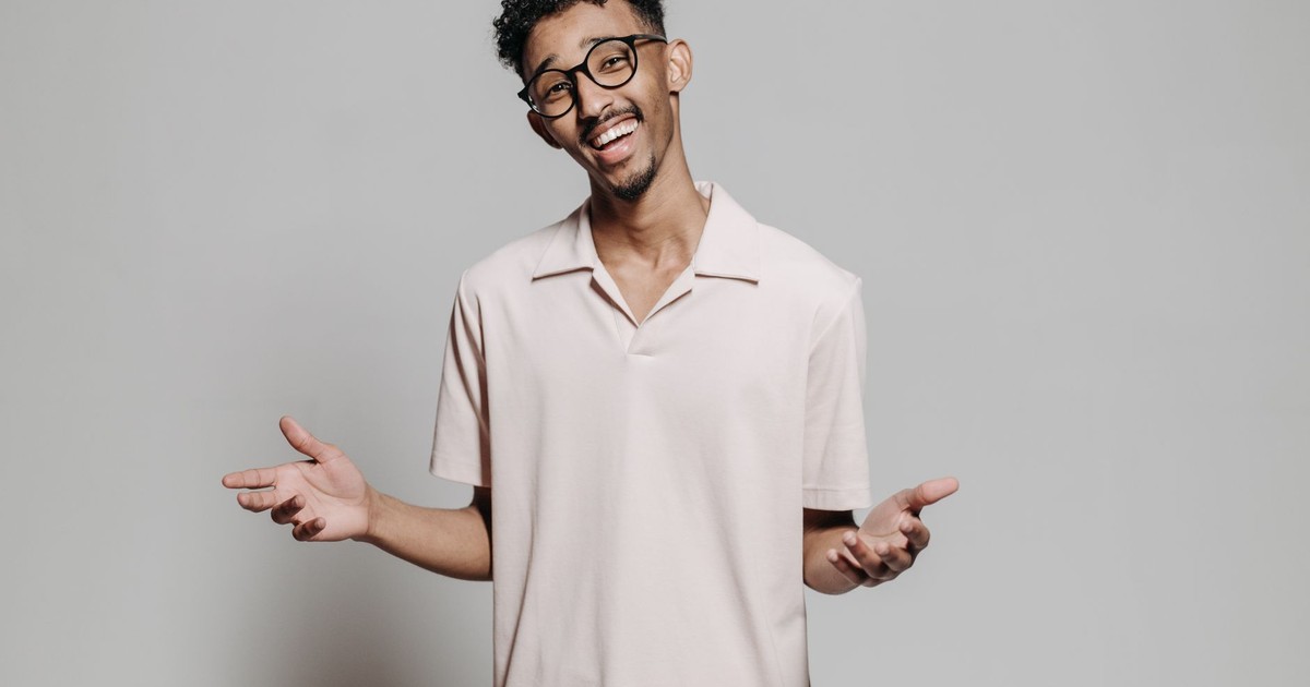 A cheerful young man wearing glasses poses against a gray studio background.
