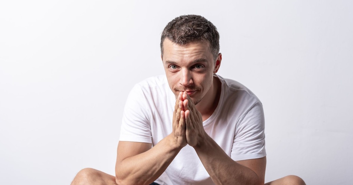 A man in a white t-shirt sits indoors with praying hands against a plain background.