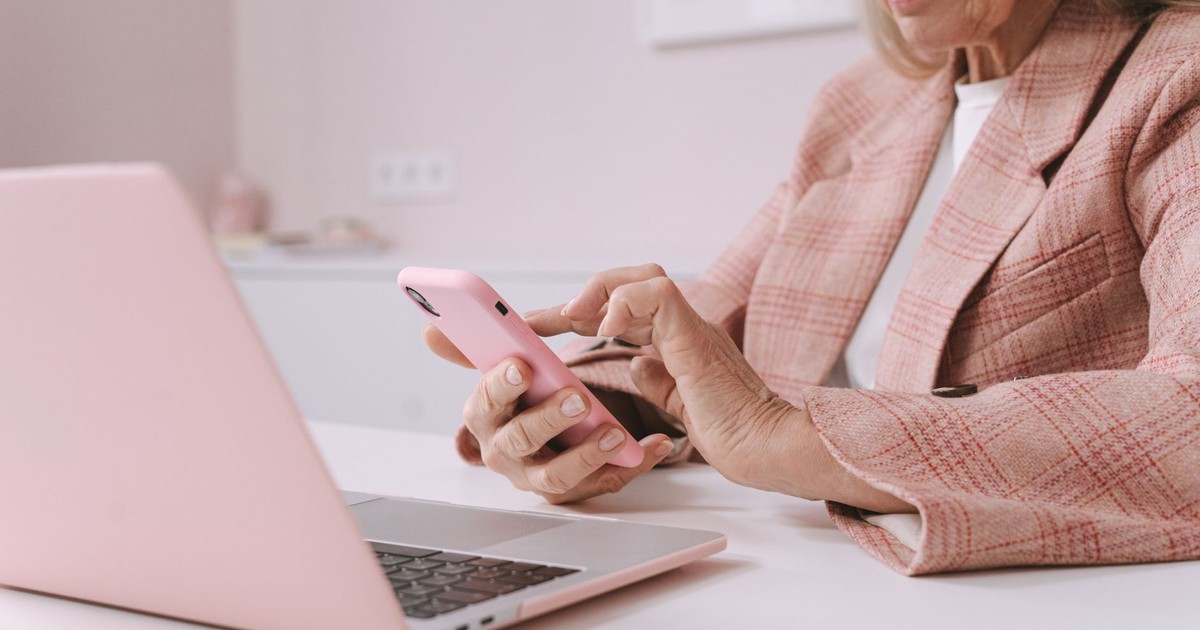 Elderly woman in pink plaid suit using a smartphone and laptop at a desk.
