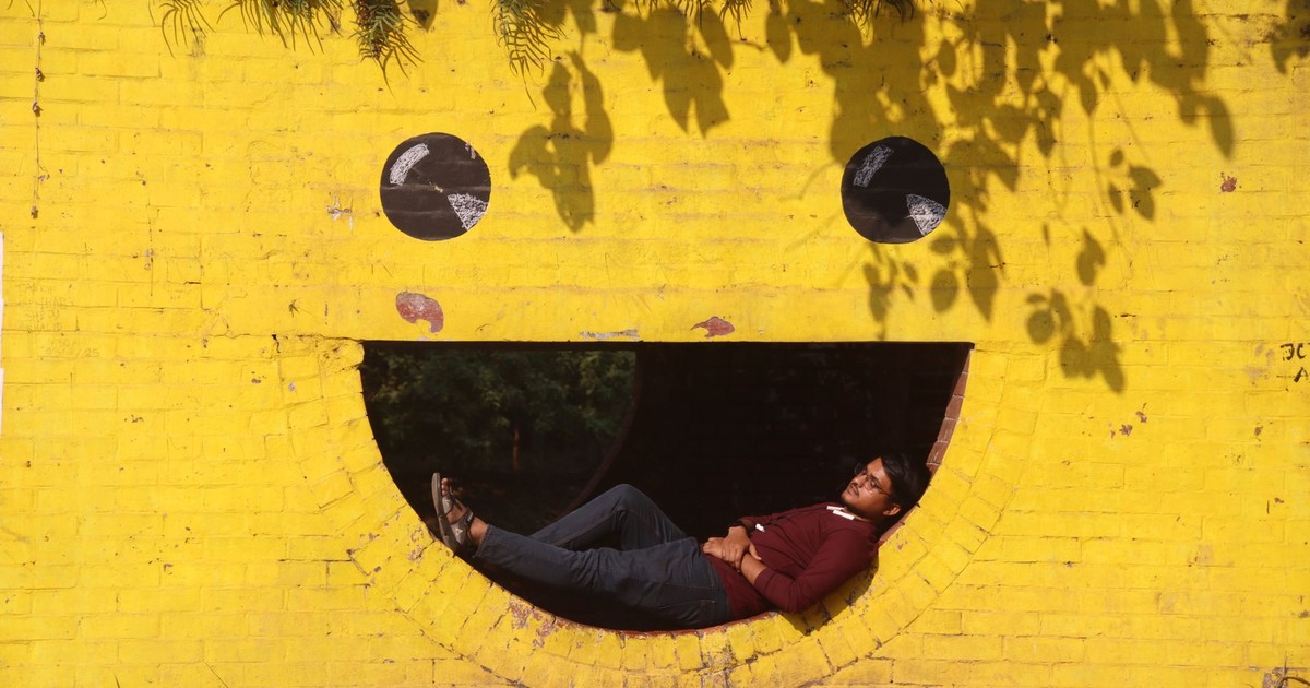 A man rests inside a smiley face cutout on a bright yellow wall under sunlit shadows.