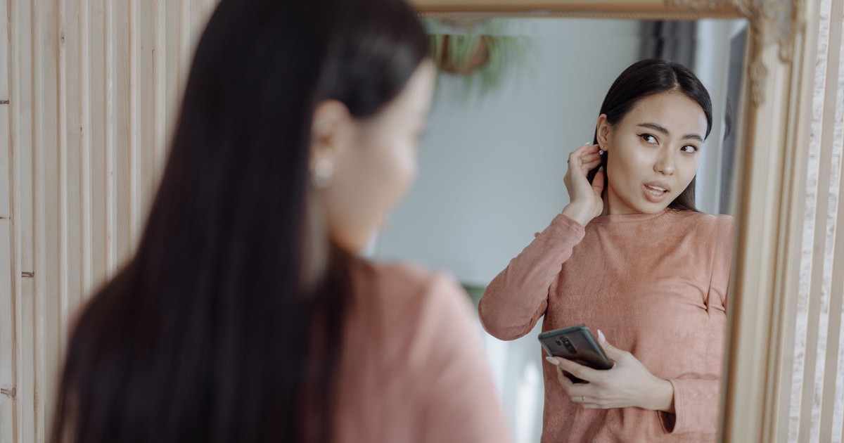 Asian woman reflects in mirror with smartphone wearing loungewear indoors.