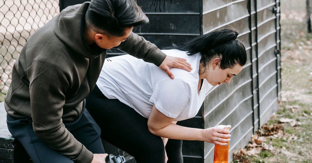 Sportsman clapping shoulder of plump tired female trainee sitting near net fence with bottle of water after training in autumn park
