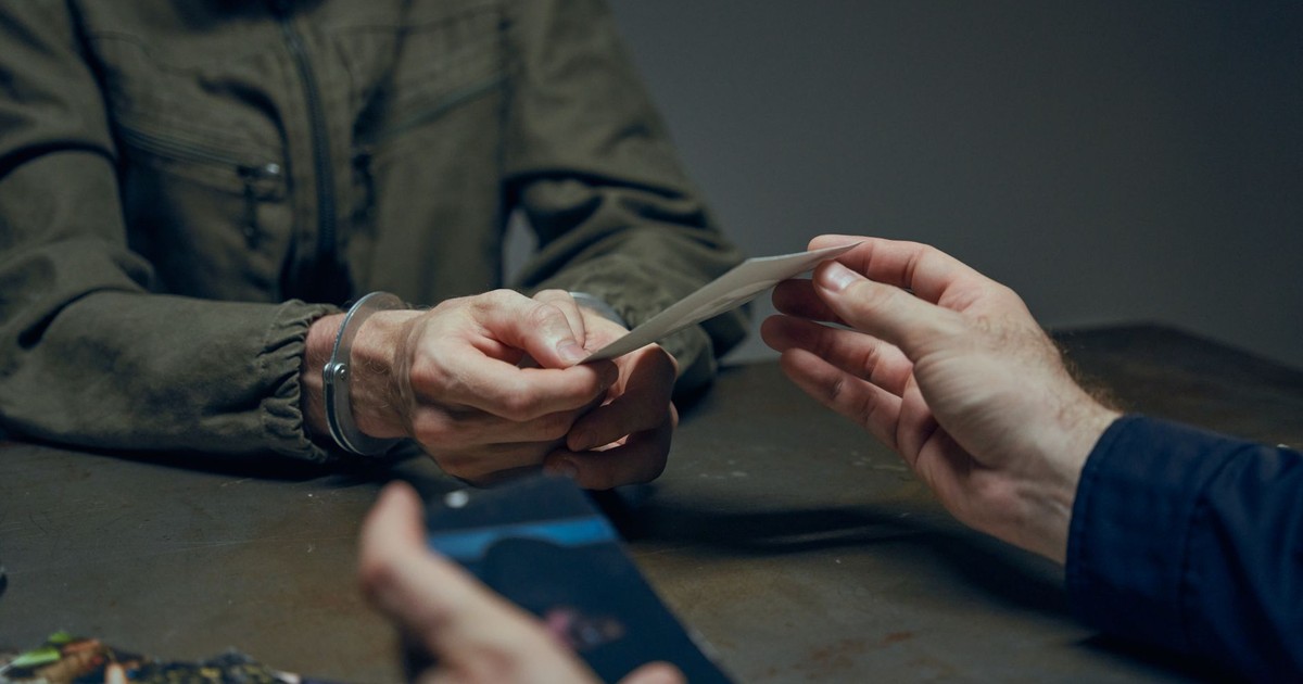 Hands exchanging evidence during an interrogation scene in a dimly lit room.