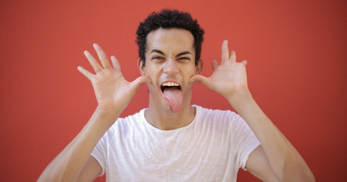 Cheerful young guy with open palms near head sticking out tongue while making grimace against red background