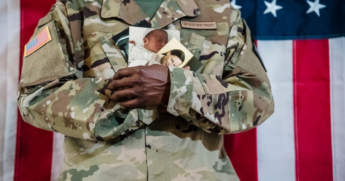 American soldier in uniform holding family photos against a flag backdrop.