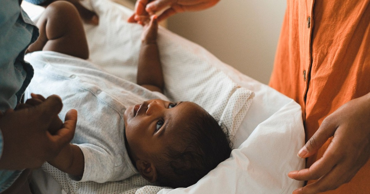 Cute baby in a gray onesie lying down, surrounded by family members' hands.