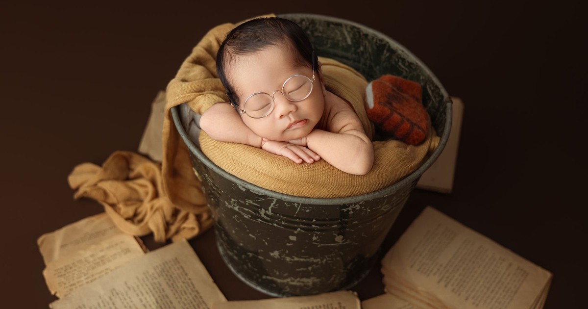 Charming newborn baby with glasses sleeping in a vintage bucket with books in a cozy setting.