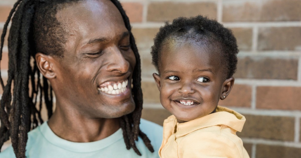A father with dreadlocks joyfully holding his smiling young daughter against a brick wall backdrop.