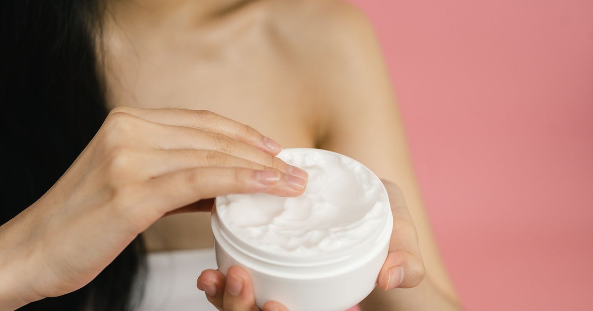 Close-up of a woman applying white cream from a jar against a pink background.