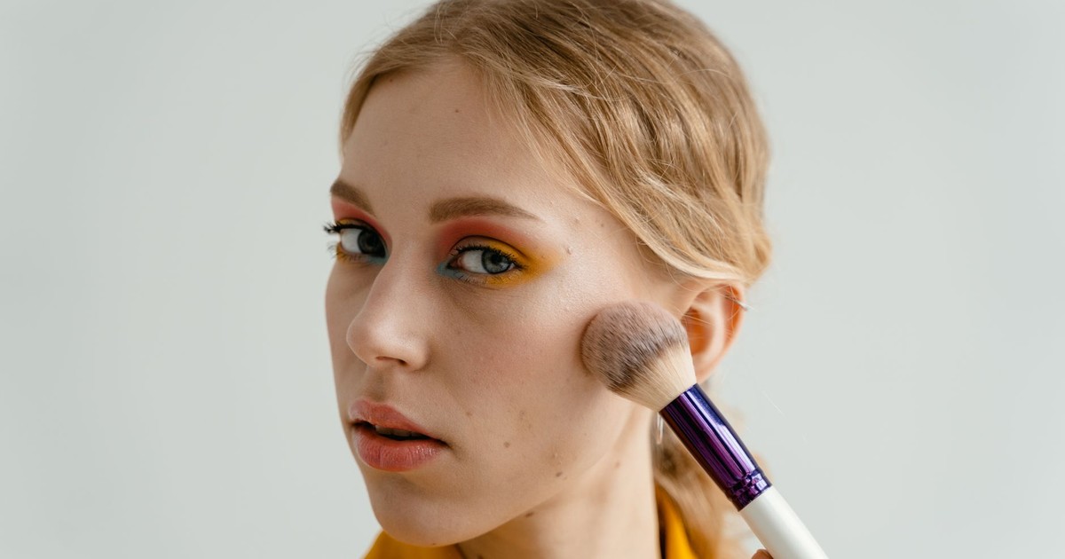 A woman applying blush with a makeup brush, showcasing a colorful eyeshadow look.