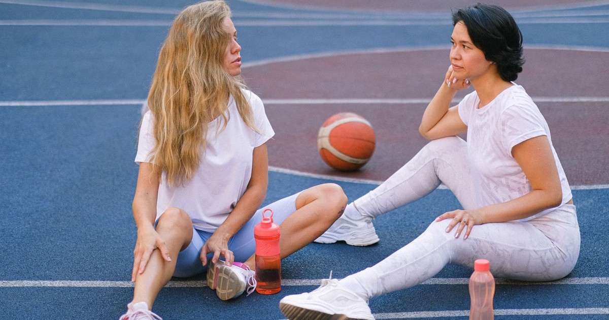 Full length of confident young ladies in sportswear speaking while having break on court after exercising with water bottles near basketball in daylight