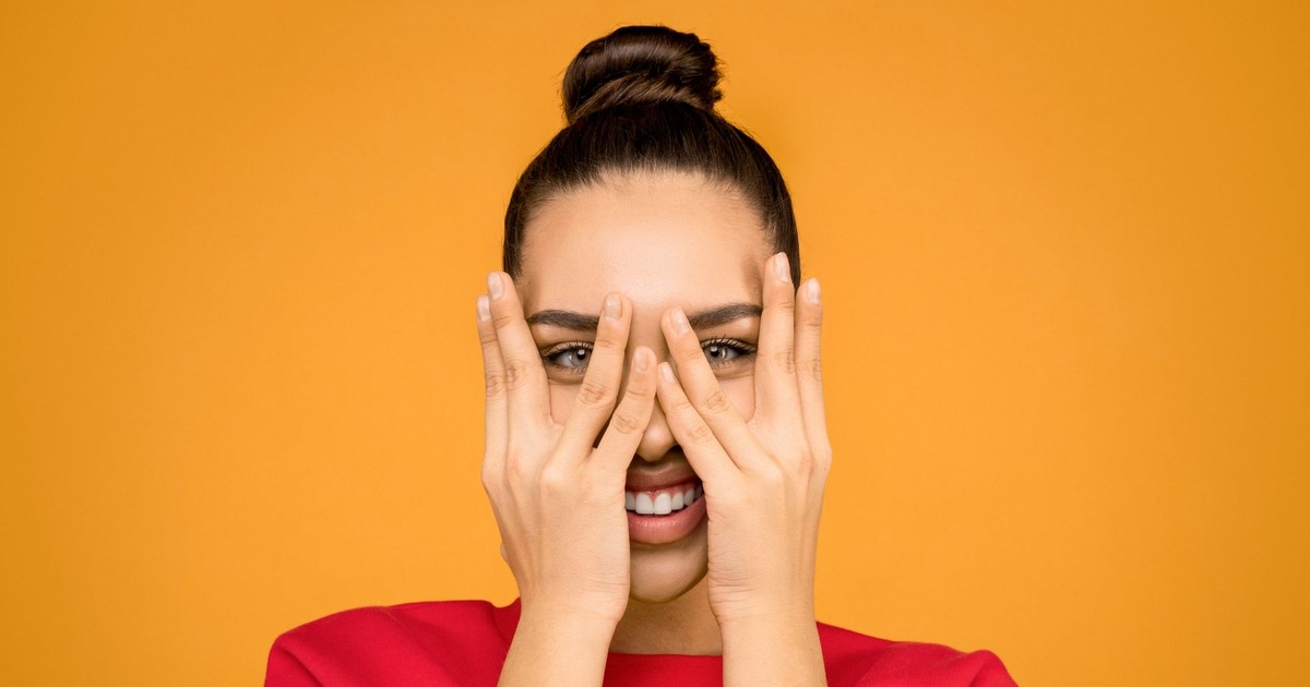 A joyful woman wearing a red sweater playfully covering her face with hands against a yellow background.