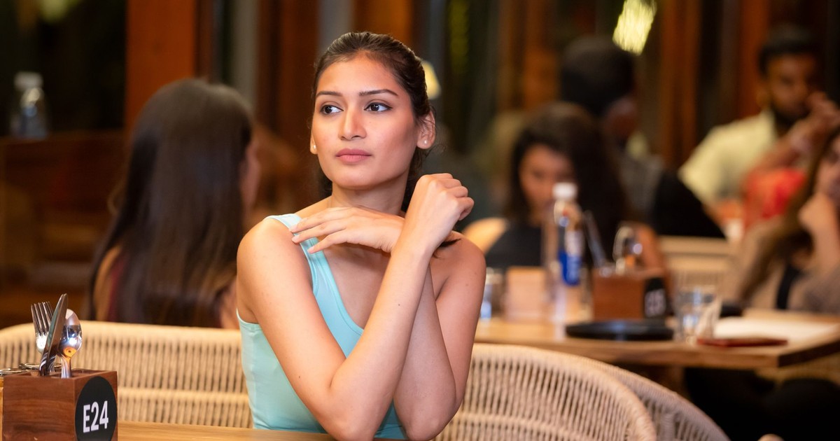A young woman in a turquoise top sits in a lively, crowded restaurant, appearing thoughtful.