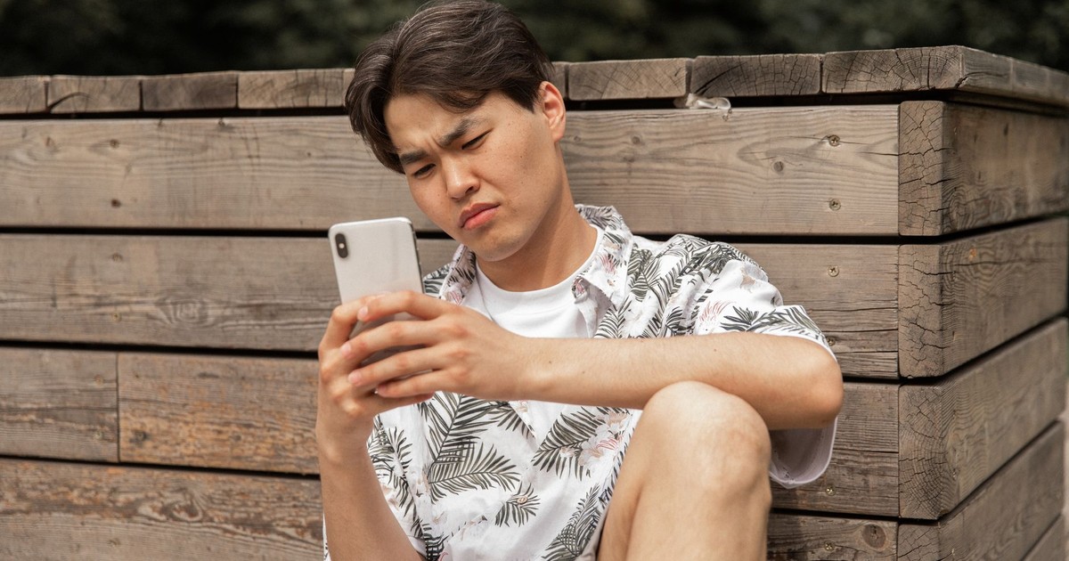 Young ethnic male in stylish clothes leaning on wooden barrier while messaging on mobile phone in park