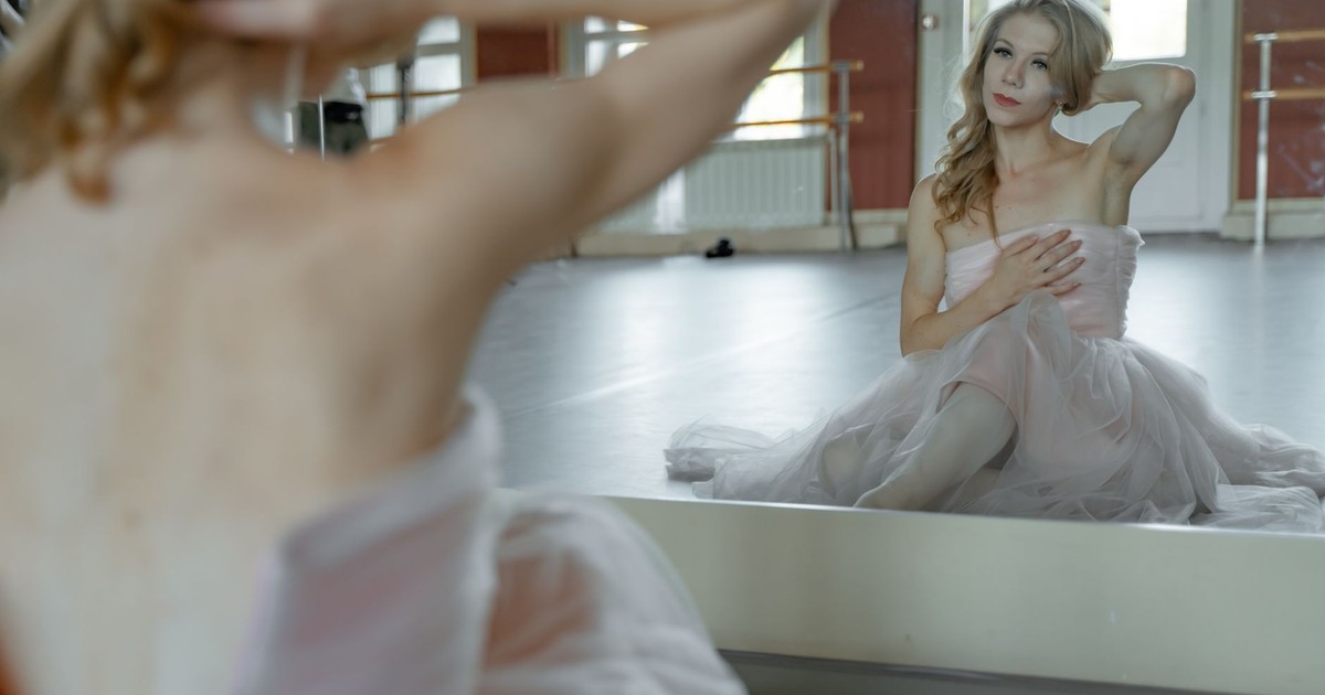 A ballet dancer in a pink dress poses in front of a mirror in a dance studio.