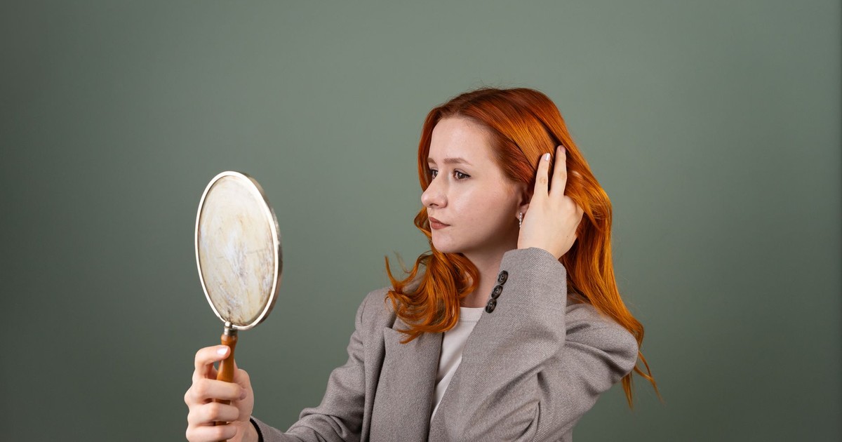 Portrait of a young woman with red hair holding a mirror, wearing a gray blazer against a green background.