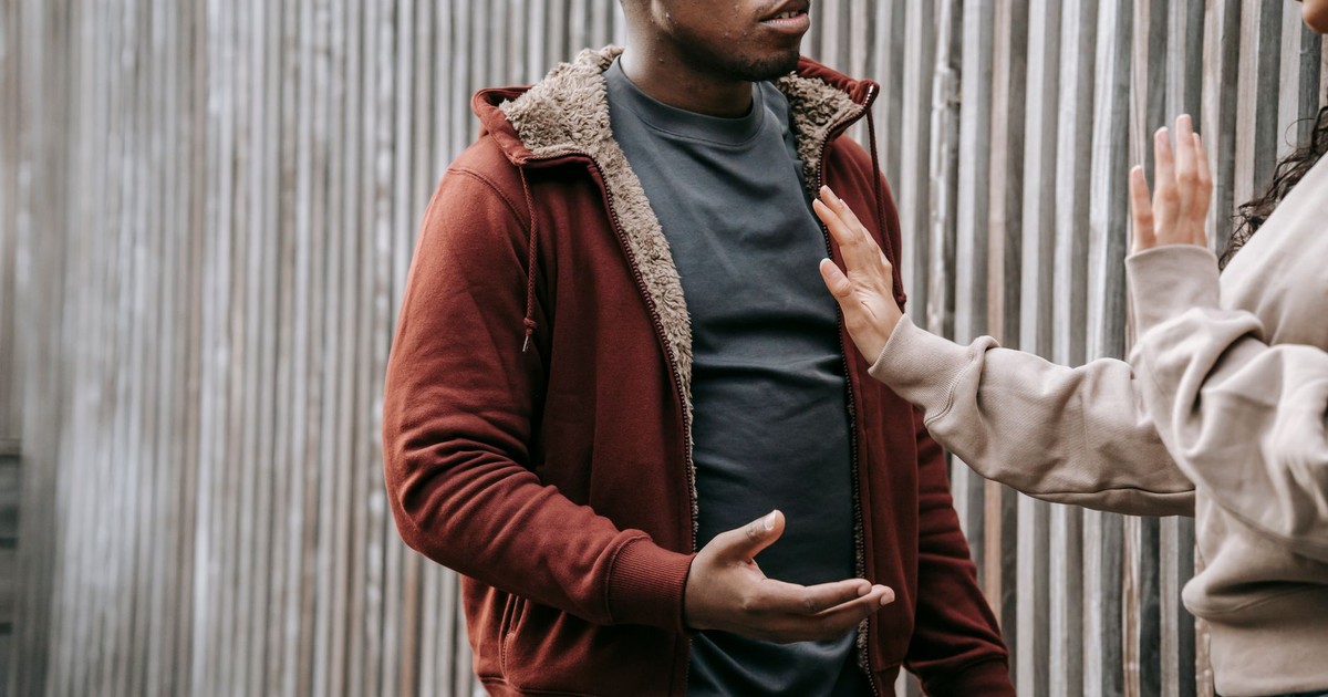 Unrecognizable woman with outstretched arm trying to stop irritated African American man while having argument on street near wooden fence