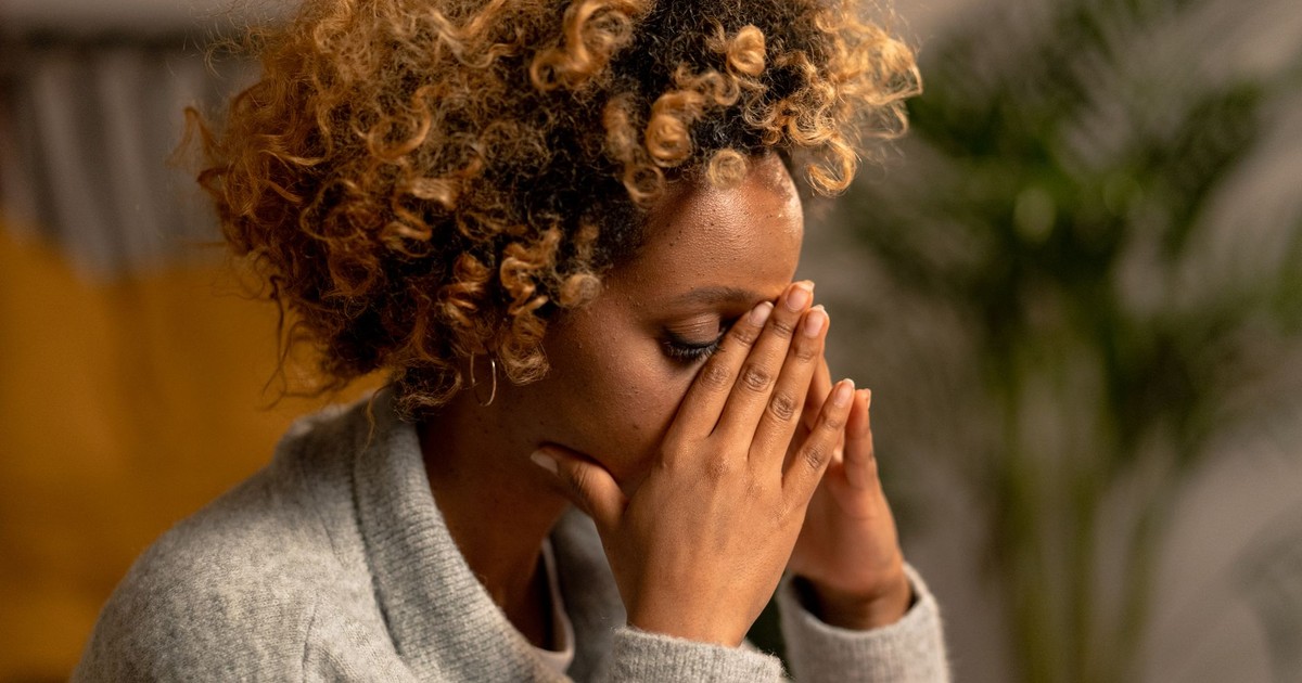 A woman with curly hair in a gray sweater, holding her face, depicting stress indoors.