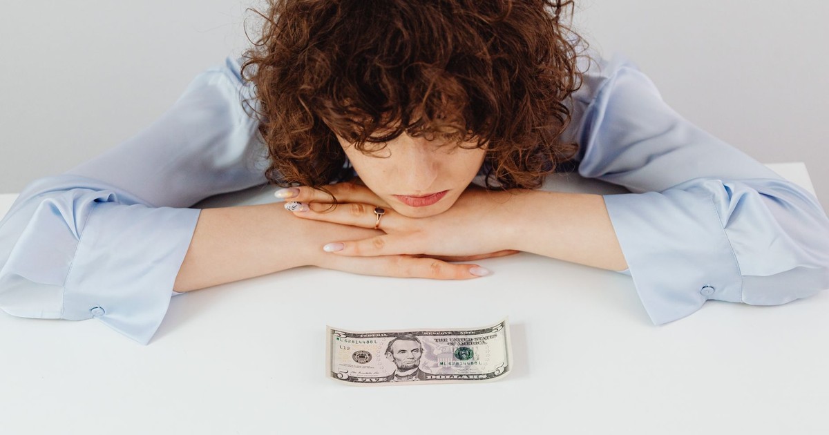 Caucasian woman with curly hair pondering over a five-dollar bill, expressing financial worry.