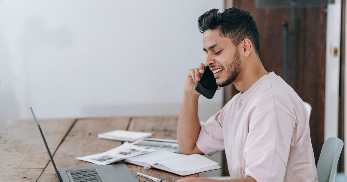 Side view of positive young Hispanic male entrepreneur in casual clothes checking documents and talking on smartphone while working remotely with laptop in home office