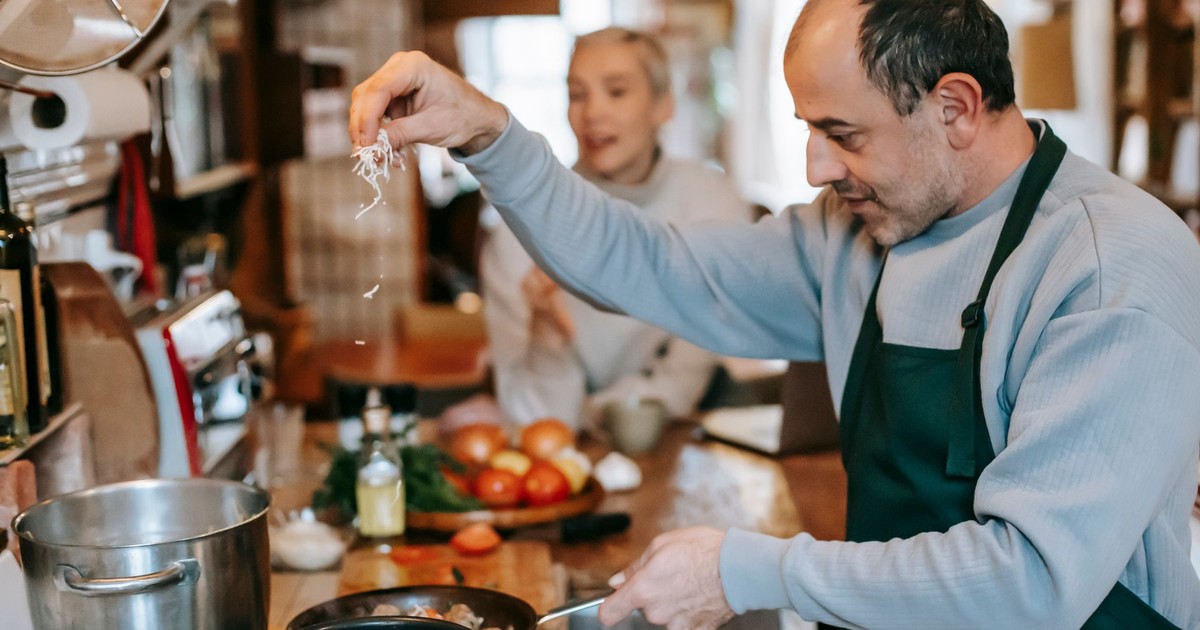 Focused male in apron adding ingredient into frying pan heating on gas stove while cooking meal for woman