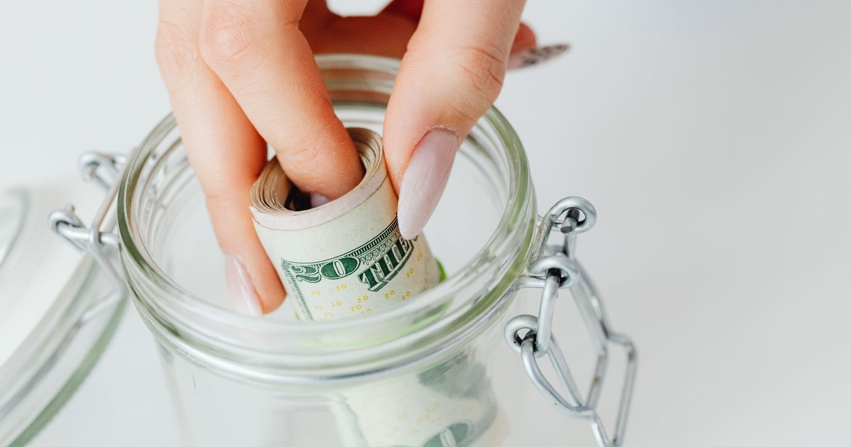 A close-up of a hand placing rolled dollars into a glass jar, symbolizing savings.