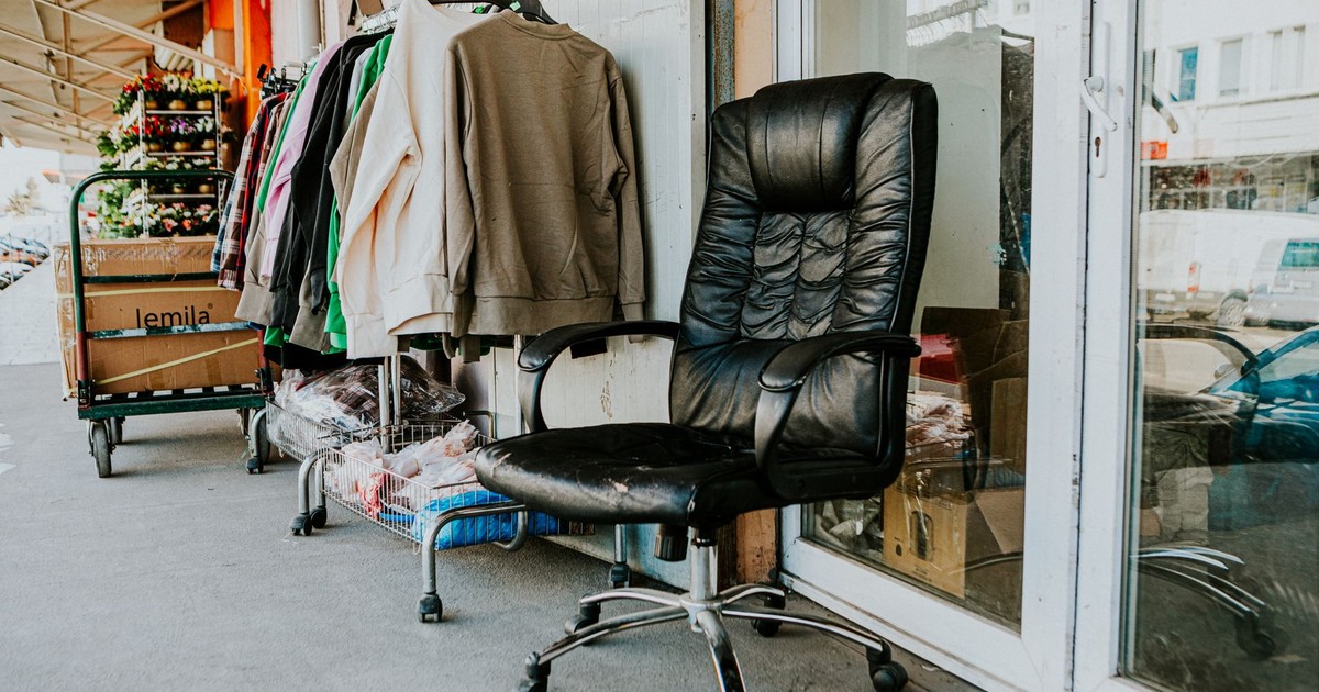 Worn black office chair outside a secondhand store with clothes and boxes nearby.