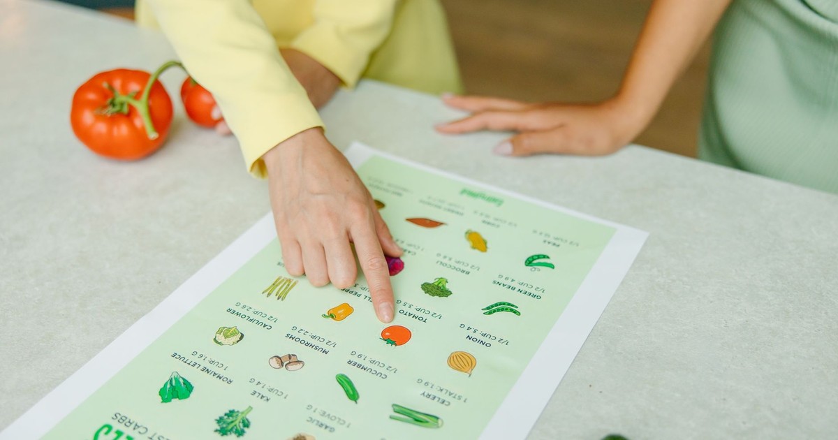 Close-up of hands pointing to a vegetable nutrition chart with fresh tomatoes on the table.