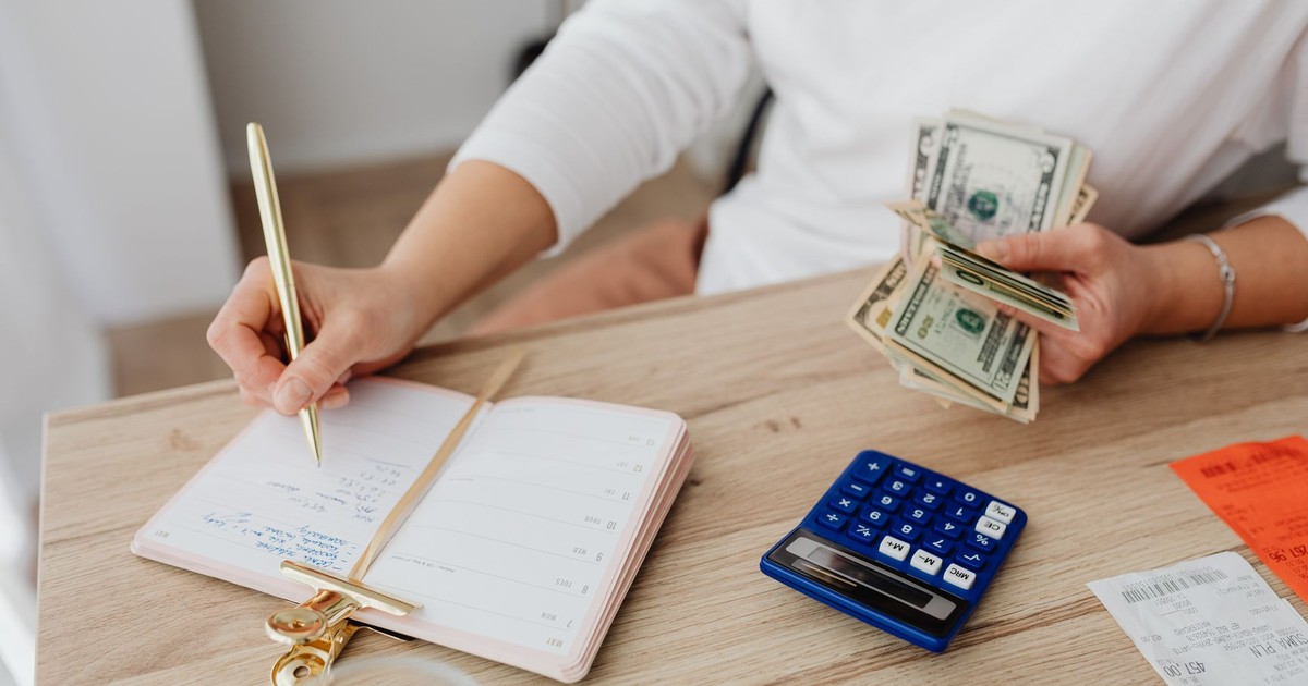 Adult holding cash and writing in planner while using a calculator at home.