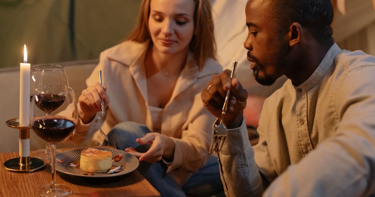 A couple enjoys a romantic candlelit dinner indoors, sharing wine and dessert.
