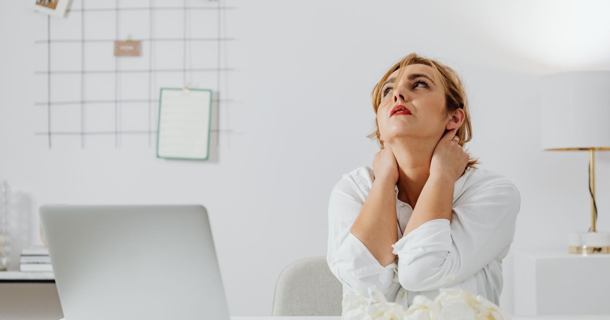 A stressed woman in white long sleeves sits at a desk with a laptop, looking upwards in an office.