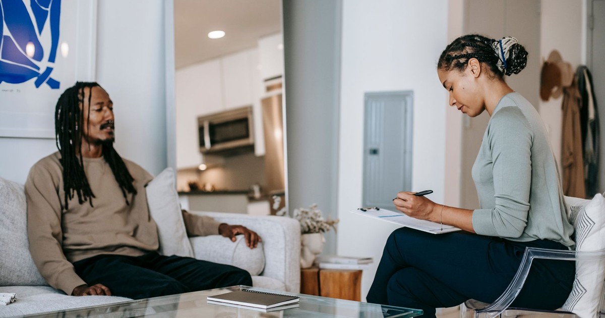 Focused African American female therapist writing on clipboard while sitting in front of upset black male client during conversation on blurred background