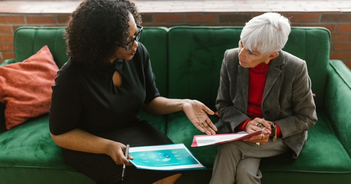 Two women in a business meeting discuss documents while sitting on a green sofa.