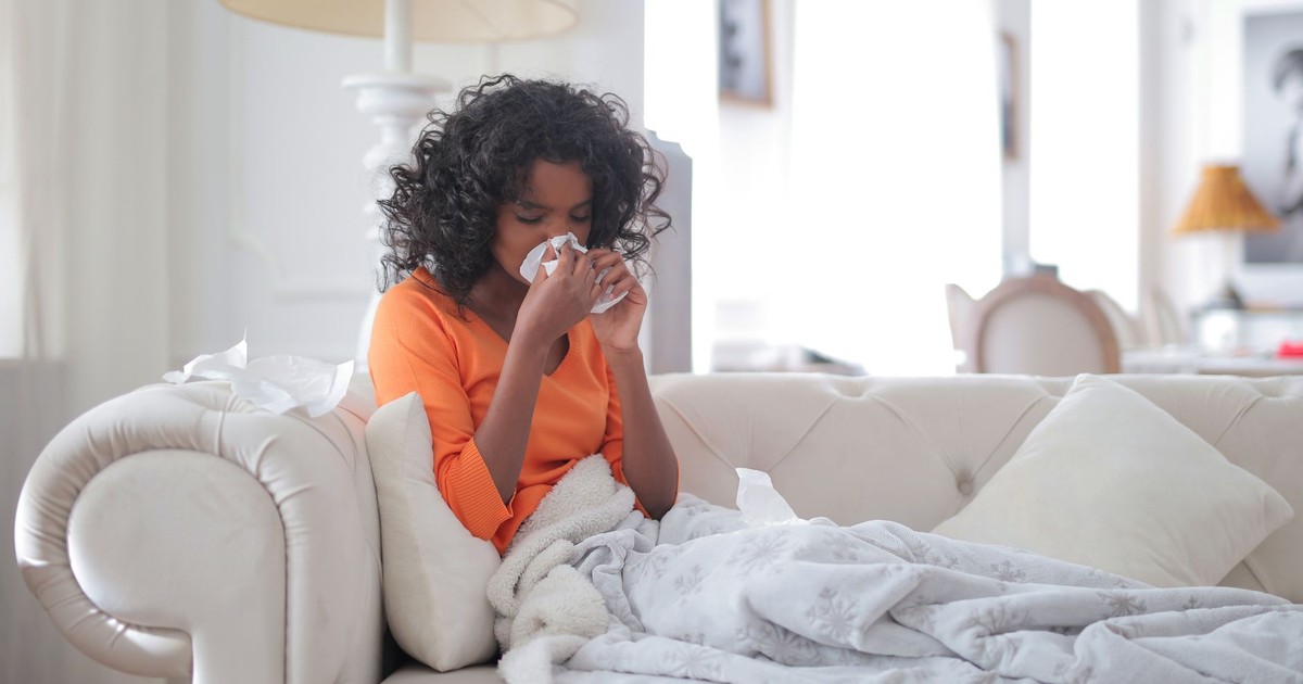 Side view of sick African American female blowing nose while sitting on sofa under blanket in cozy modern home in daylight