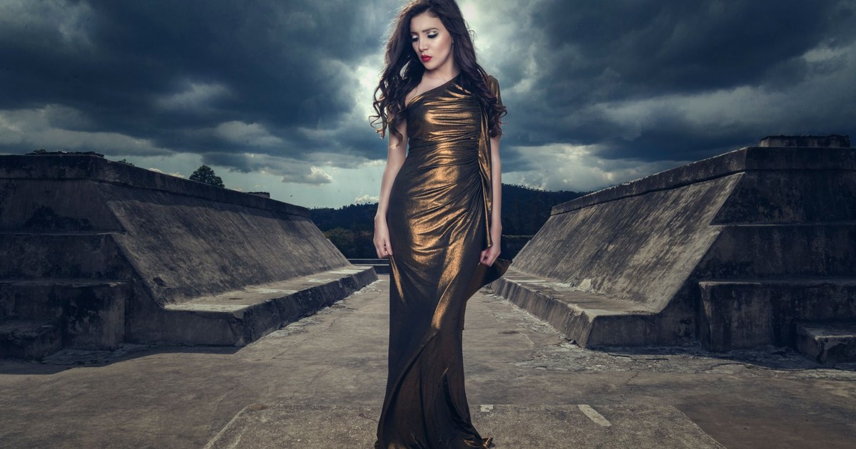 A woman in a luxurious dress poses outdoor amidst concrete structures under a dramatic sky.