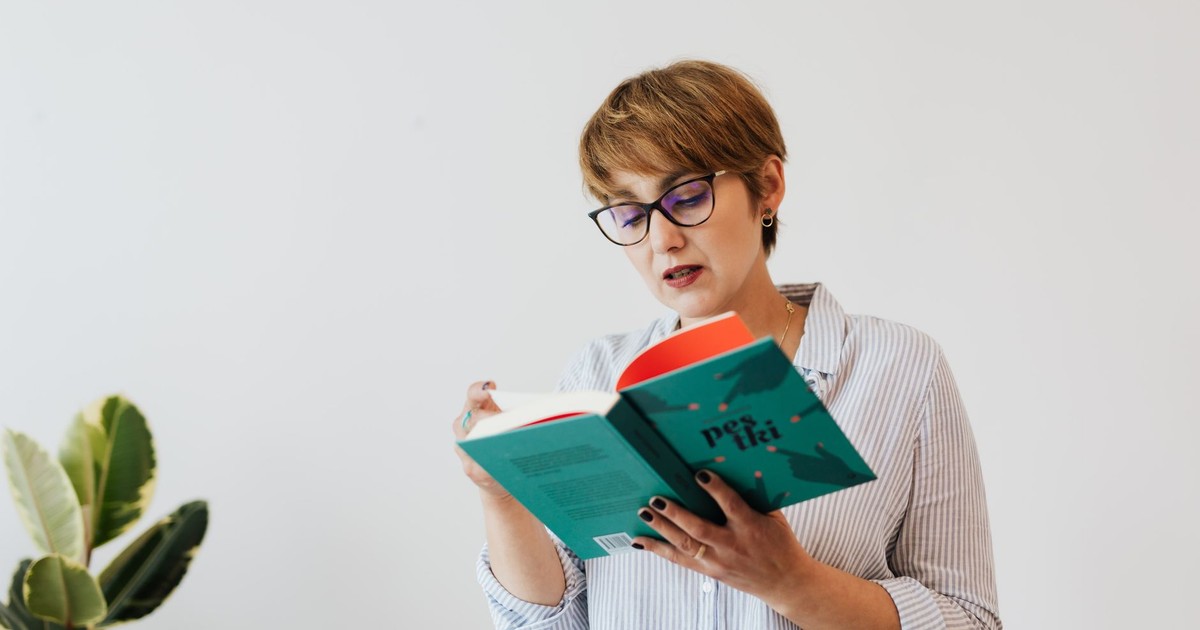 Concentrated female in casual shirt and eyeglasses reading interesting book with green hardcover while standing against white wall and houseplant