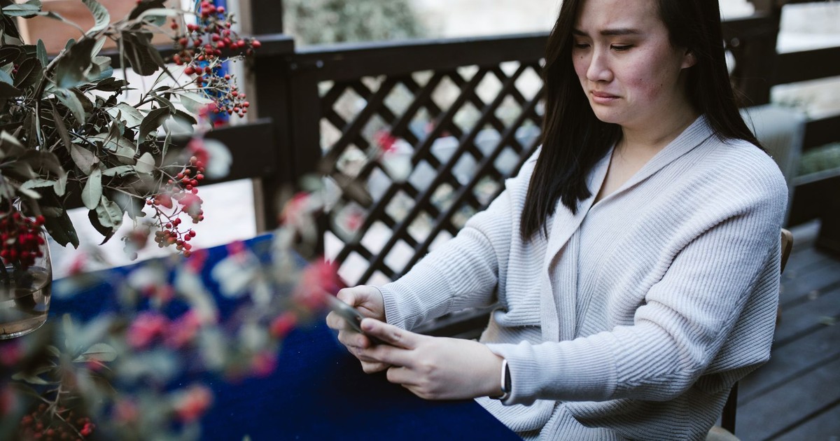 A woman appears upset while sitting outdoors using her phone, surrounded by decorated branches.
