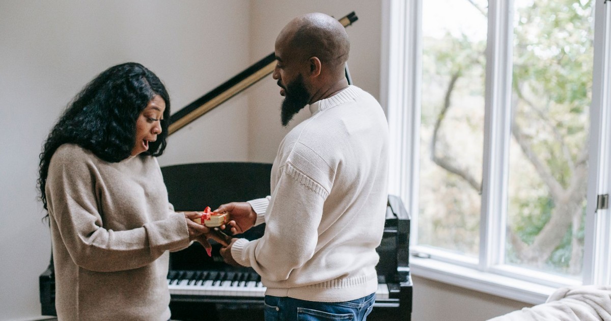 Side view of surprised African American woman receiving present from beaded husband during holiday celebration in room with piano near window