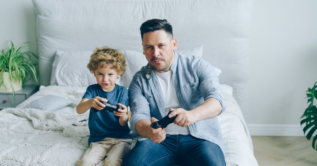 Father and son joyfully playing video games while sitting on a bed, capturing a moment of family fun.