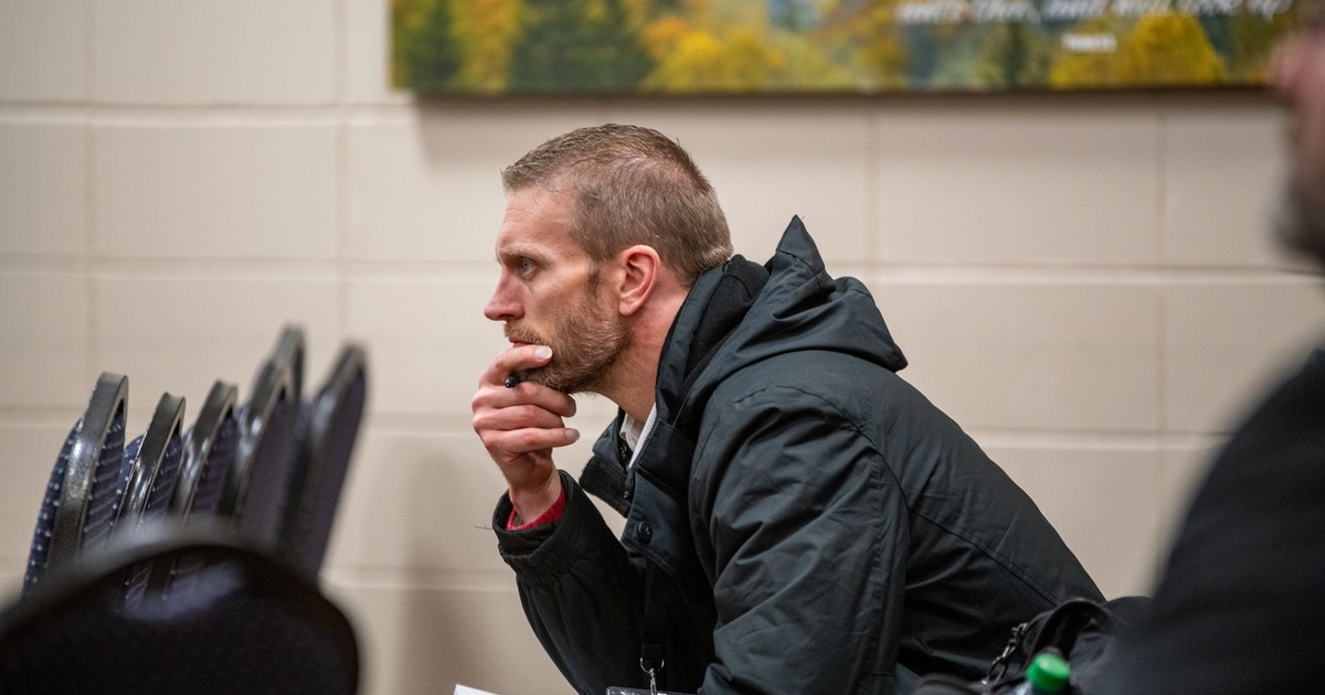 Man in a black jacket sits thoughtfully indoors, hand on chin, contemplating.