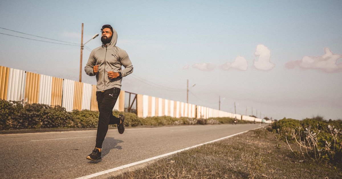 A bearded man in a hoodie jogs along an empty road under a clear sky.