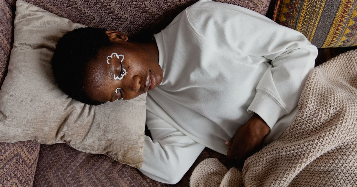 An adult woman relaxing indoors with artistic face paint, lying on a pillow and blanket in a cozy setting.