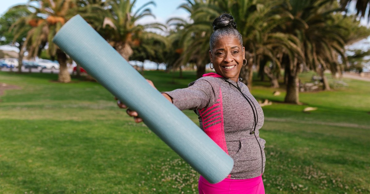 Smiling senior woman holding yoga mat, preparing for outdoor exercise in a sunny park.
