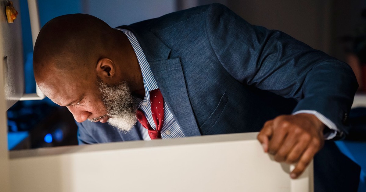 Smartly dressed man peering into a refrigerator, possibly late at night at home.