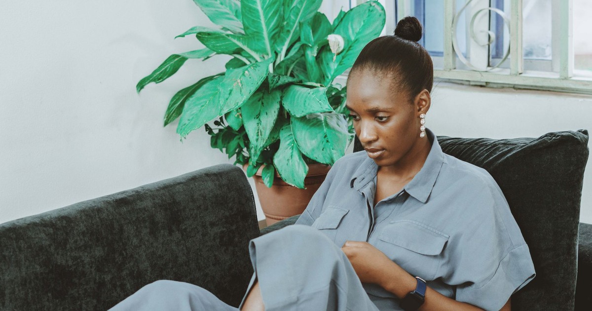 A young woman in a gray dress sits on a sofa using a smartphone, with a green plant in the background.
