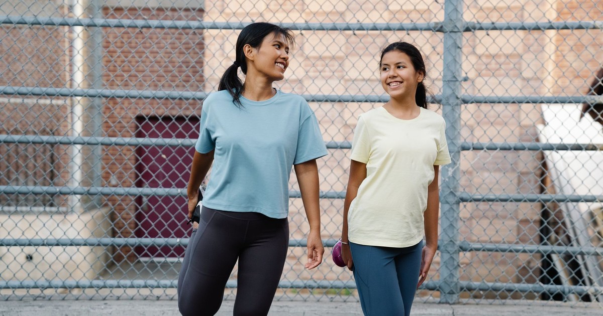 Two women in sportswear stretching outdoors beside a mesh fence, promoting fitness and health.