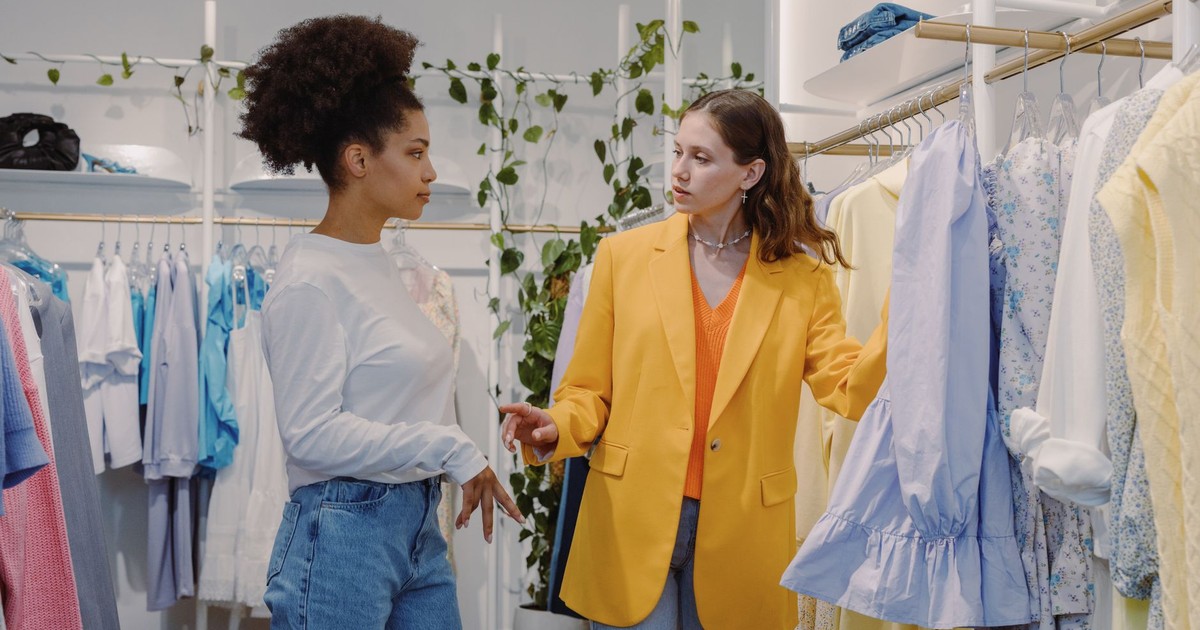 Two women in a boutique store browsing clothes, discussing styles.