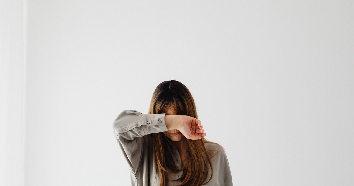 A woman with long hair covers her face with her arm against a white backdrop.